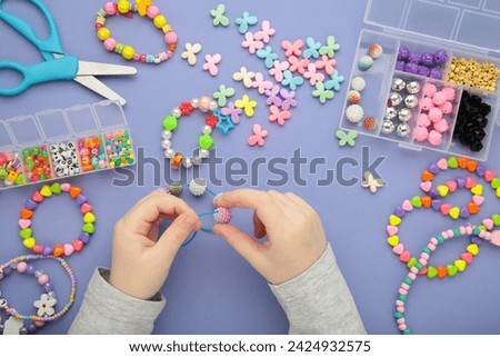 Little girl made bracelets on purple background. Kids handmade beaded jewelry. Necklaces and bracelets made from multicolored beads and pearls. DIY bracelet beads. Royalty-Free Stock Photo #2424932575
