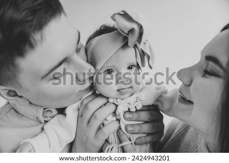 Father and mother hugging daughter closeup. Happy family hugs cute baby on holiday. Dad, mom holds hands infant. Daddy, mommy embrace girl with bow in hair isolated on wall. Black and white photo.