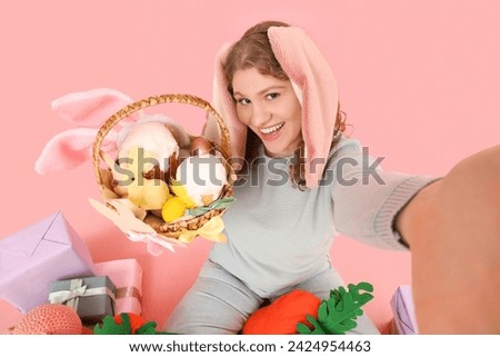 Beautiful young happy woman in bunny ears with carrot-shaped toy, gift boxes and Easter basket taking selfie on pink background