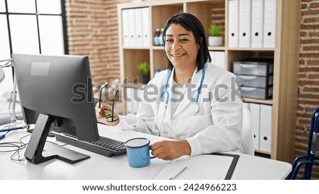 A smiling hispanic woman doctor sits at a desk with a computer in a clinic office setting.