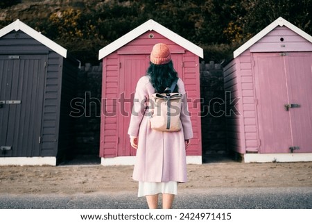 Back view stylish hipster woman with color hair in total pink outfit and backpack looking at wooden beach huts. Off season Travel concept. Seasonal street fashion. Barbiecore style. Simple pleasures. Royalty-Free Stock Photo #2424971415