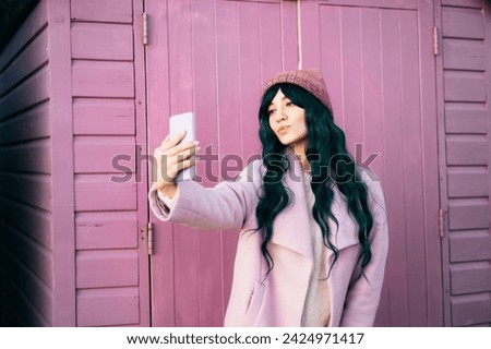 Stylish young hipster woman with color hair wearing pink coat, knitted hat taking a selfie photo on phone on pink beach hunt background. Blogger, influencer. Seasonal fashion, Barbiecore style trend. Royalty-Free Stock Photo #2424971417