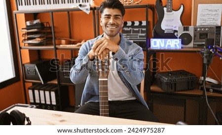 A smiling young hispanic man with a beard in a music studio, holding a guitar, sitting in front of a microphone and 'on air' sign.