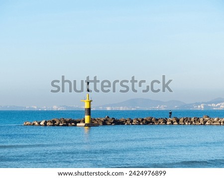 Coastal beacon in breakwater on a sunny day with blue sky. Man fishing and mountains in the background