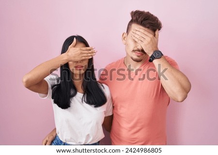 Young hispanic couple standing over pink background covering eyes with hand, looking serious and sad. sightless, hiding and rejection concept  Royalty-Free Stock Photo #2424986805