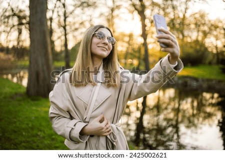 Stylish beautiful young girl in sunglasses and a beige trench coat takes a selfie against the backdrop of trees and a river lake
