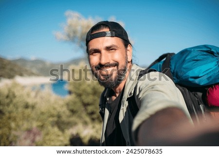 A male tourist with a large backpack and hiking gear takes a selfie.  A young satisfied guy smiles, shoots himself on camera on a walking trip