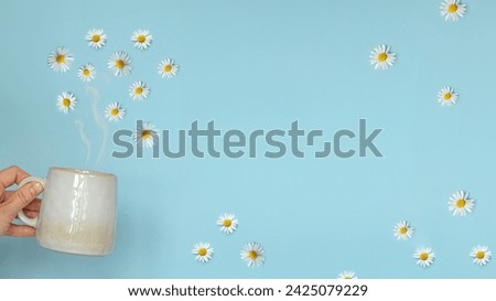 Persons hand holding a steaming cup of hot chamomile tea, with delicate white flowers floating on the surface. The background is a soothing light blue, creating a calm and serene atmosphere.