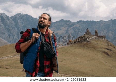 A male tourist on the background of the Trinity Church in Gergeti in the mountains on a clear day. A young guy and an Orthodox church, a chapel on a hill against the background of mountains.