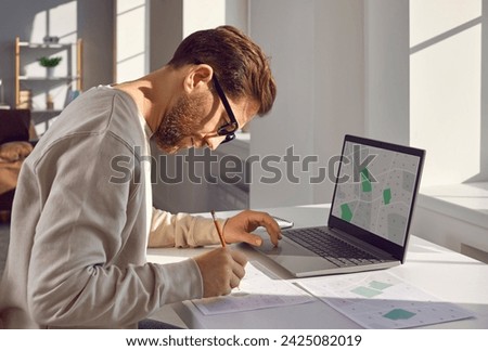 Professional cartographer in glasses working at home or at office with printed cadastral map on his workplace. Young man in casual clothes analyzing cadastral map and searching for a building plot. Royalty-Free Stock Photo #2425082019