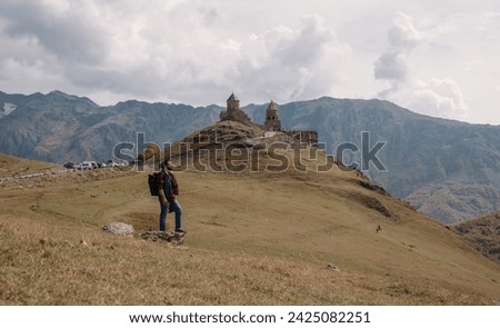 A male tourist on the background of the Trinity Church in Gergeti in the mountains on a clear day. A young guy and an Orthodox church, a chapel on a hill against the background of mountains.