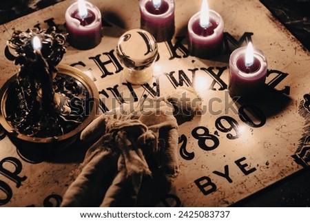 A dark atmospheric setting with an Ouija board, candles, and mystical objects invoking a sense of the occult. Royalty-Free Stock Photo #2425083737
