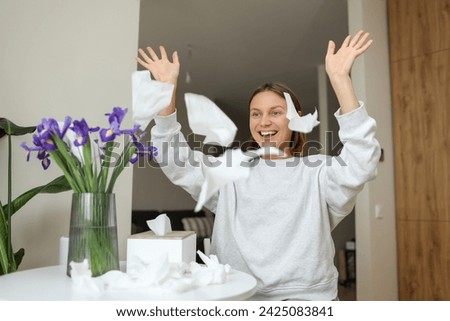 Seasonal allergy free. Happy young woman throws tissues up at home. Healthy life without of runny nose, itching or cough symptoms. Allergen bouquet of iris flowers at vase and used napkins on table Royalty-Free Stock Photo #2425083841