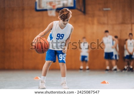 Dynamic portrait of a junior basketball player dribbling a ball and practicing moves at indoor court on training. In a blurry background are his teammates waiting for their turn. A boy with basketball