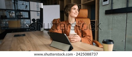 Portrait of young confident woman, employer in casual clothes, working in office, has digital tablet and coffee on tablet, smiling with self-assured face expression.