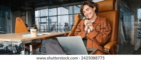 Portrait of woman in office, looking delighted, wearing wireless earphones for online meeting, joins online chat, looks at laptop.