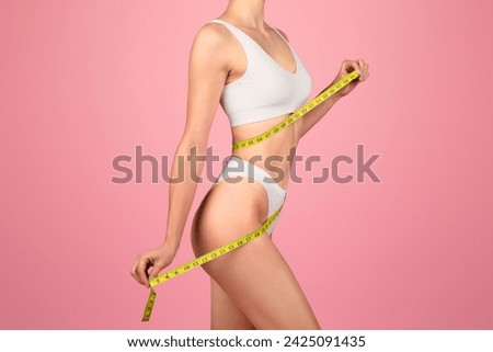 Side profile of a fit woman measuring her waist with a yellow tape measure, dressed in a white sports bra and underwear, against a vibrant pink background, showcasing health and body consciousness Royalty-Free Stock Photo #2425091435
