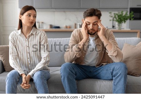 Upset young man touching head, showing signs of stress, while concerned woman looking at husband, highlighting relationship tension Royalty-Free Stock Photo #2425091447