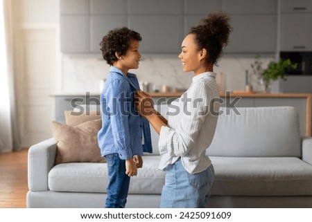 Affectionate young black mother and her curly-haired son making eye contact, holding hands, with expressions of love and trust in bright living room, side view Royalty-Free Stock Photo #2425091609