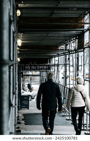 A pair of seniors walking down the sidewalk of Burrard Street, under a scaffolding raised for construction work. An overhanging sign advertises a coffee shop hidden by the scaffolding is still open.
