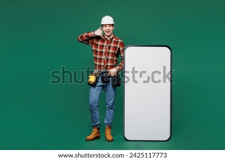 Full body young laborer man wear red shirt hardhat big huge blank screen area mobile cell phone do call back gesture isolated on plain green background. Instruments for renovation. Repair home concept