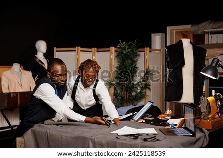 High end manufacturing suitmakers cutting textile material with scissors in tailoring studio, designing sartorial fashion clothes. Dressmakers working on bespoke client comission Royalty-Free Stock Photo #2425118539