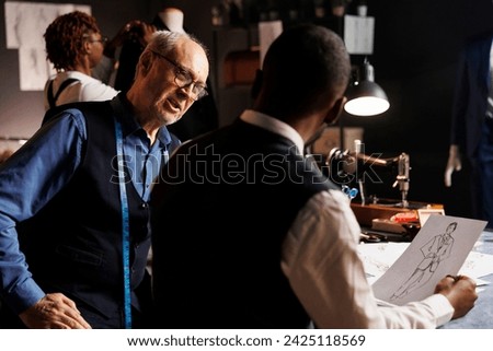 Couturiers manufacturing bespoken clothing, working together to finalize client sartorial comission. Master tailor and diverse apprentice team of suitmakers checking fashion design sketches in atelier