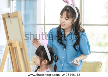 Portrait shot of smiling young asian woman pretty mother helping little cute daughter girl paint picture on canvas having fun together and spending family leisure time in living room at home.