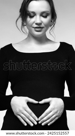 Black and white portrait of young pretty pregnant woman on gray studio background. Female in black dress with hands near pregnant belly with heart sign.