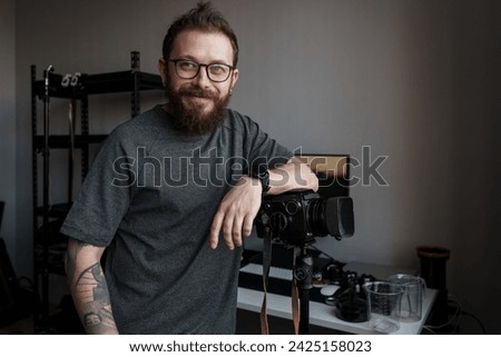 A confident photographer stands by his medium format camera mounted on a tripod, poised to capture images in his well-equipped studio