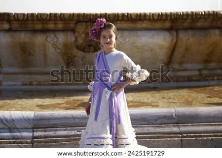 a pretty little girl dancing flamenco dressed in a beige dress with ruffles and purple fringes in a famous square in seville, spain. The girl has a flower in her hair. In the background a big fountain