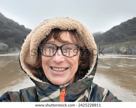 Mature woman wears a camouflage robe with the hood up to keep warm while walking on the beach in the rain. Her glasses are wet and she takes a selfie for social media.