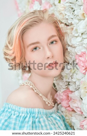 A charming blonde teenage girl with a short haircut in a summer sundress and a pearl necklace poses against a wall of white and pink flowers. Delicate spring-summer look. Beauty, cosmetics.