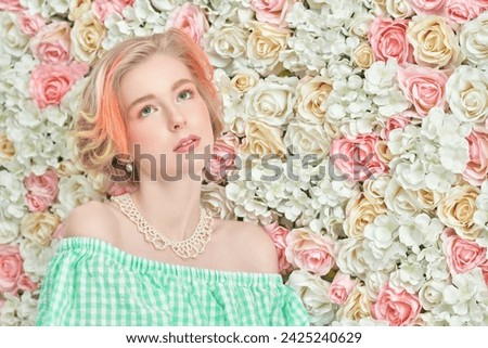 Beauty, cosmetics. A cute blonde teenage girl with a short haircut in a summer sundress and a pearl necklace poses against a wall of white and pink flowers. Delicate spring-summer look. Copy space.
