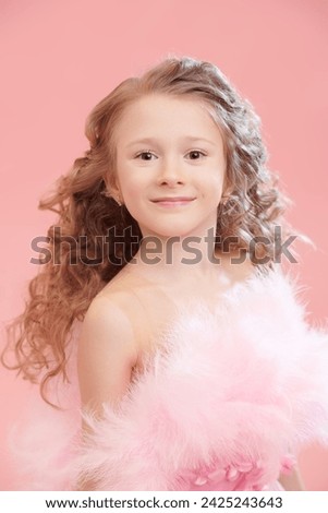 Portrait of a charming little girl with lush curly hair and in a pink evening dress with feathers smiling. Pink studio background. Children's holiday fashion. Little Princess.