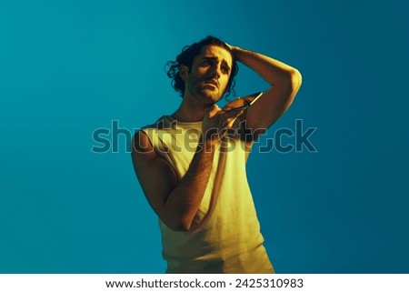 Young man with curly hair and unshaved face, in casual clothes recording voice message on mobile phone against blue background in neon light. Concept of youth, human emotions, lifestyle