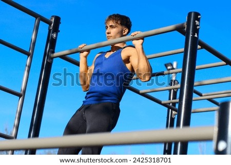 Caucasian young man doing pull-ups in an outdoor bars park, dressed in sportswear, with blue sky in background. Concept of workout and calisthenics