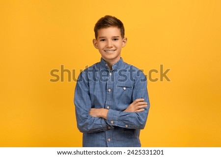 Confident teen boy in blue denim shirt standing with arms crossed, smiling gently while posing against bright yellow background, male kid depicting confidence while looking at camera