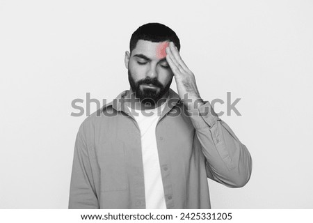 Unhappy young latin man with beard in casual put hand on forehead, suffer from headache, inflammation red, isolated on gray background, studio. Stress, health problems, migraine, black and white Royalty-Free Stock Photo #2425331205