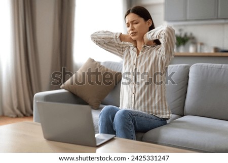 Tired young woman massaging her neck, showing signs of strain and discomfort while working from home, sitting in front of her laptop on sofa