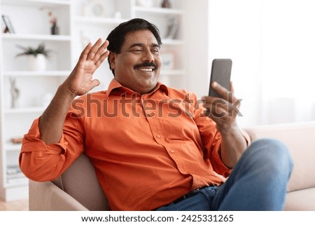 Cheerful mature indian man with moustache siting on couch at home, waving at cell phone screen, have video call with friend or lover, copy space. Telecommunication concept, mobile app