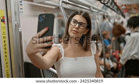 Beautiful hispanic woman charmingly smiling, standing in the bustling subway, taking a radiant selfie with her glasses on the urban city underground.