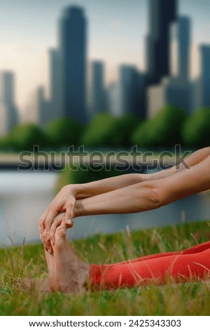 Closeup photo of barefoot sporty girl practicing stretching yoga pose during training while sitting on grass against urban background