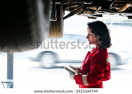 A Caucasian female mechanic in a red uniform standing under the car's bottom and checking digital tablet in the garage. A woman smiling while holding a wrench. Car repair service. Vehicle maintenance