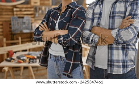 Two relaxed carpenters, a man and a woman, standing together with the bond of woodworking. their crossed arms gesture displays professional confidence at the indoor carpentry business. Royalty-Free Stock Photo #2425347461