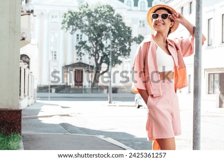 Young beautiful smiling hipster woman in trendy summer pink costume clothes. Carefree female posing in the street at sunny day. Positive model outdoors at sunset. Cheerful and happy in hat, sunglasses