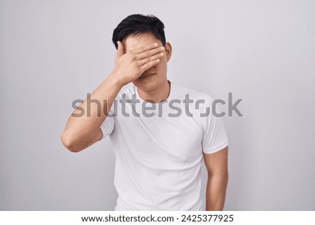 Young asian man standing over white background covering eyes with hand, looking serious and sad. sightless, hiding and rejection concept  Royalty-Free Stock Photo #2425377925