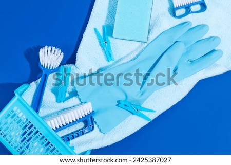 A plastic basket with cleaning supplies decorated on blue background. Brushes, rubber glove and clothespins displayed. Scene for advertising cleansing product. Creative ideas