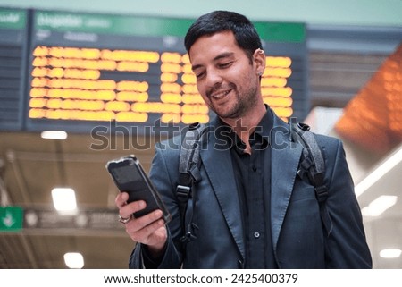 young latino man travelling the world. man looking at his smart phone in the station with the travel schedule in the background. Royalty-Free Stock Photo #2425400379