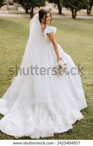 Brunette bride in a white dress with a long train, holding a bouquet and walking in the garden. Wedding photo session in nature. Beautiful hair and makeup. Celebration
